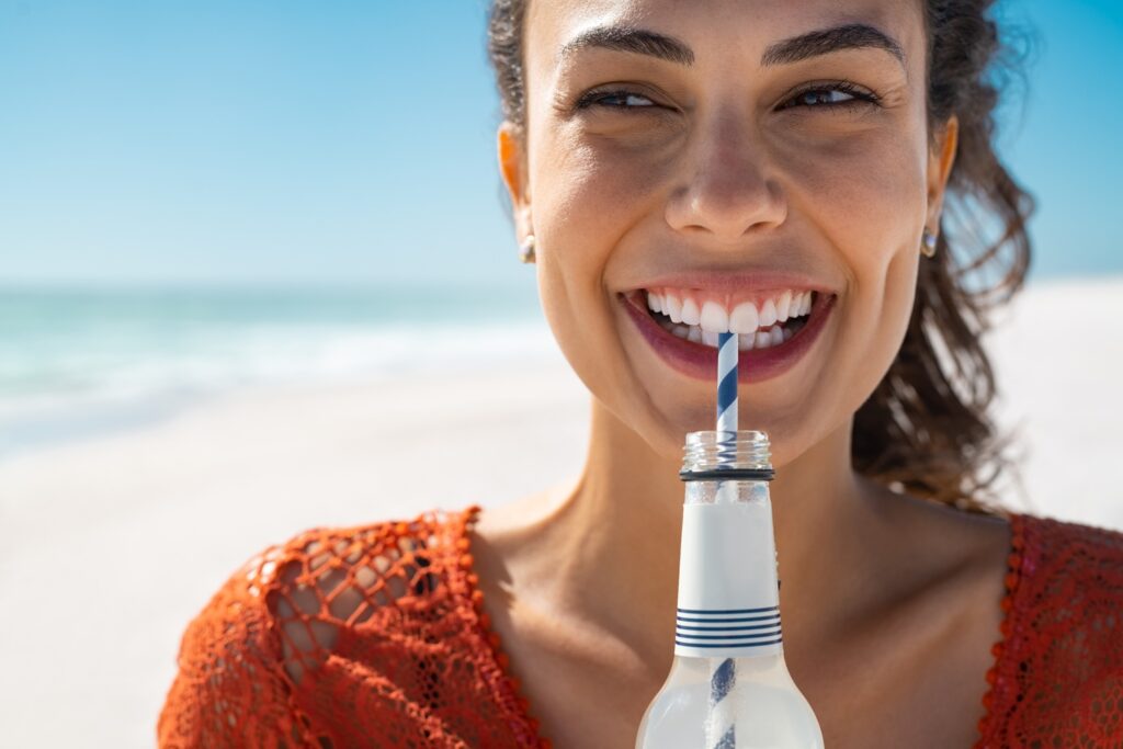 Chica en la playa sonriendo mientras bebe un refresco con pajita | Cuidado Dental | Higiene Bucal | Clínica dental Legazpi